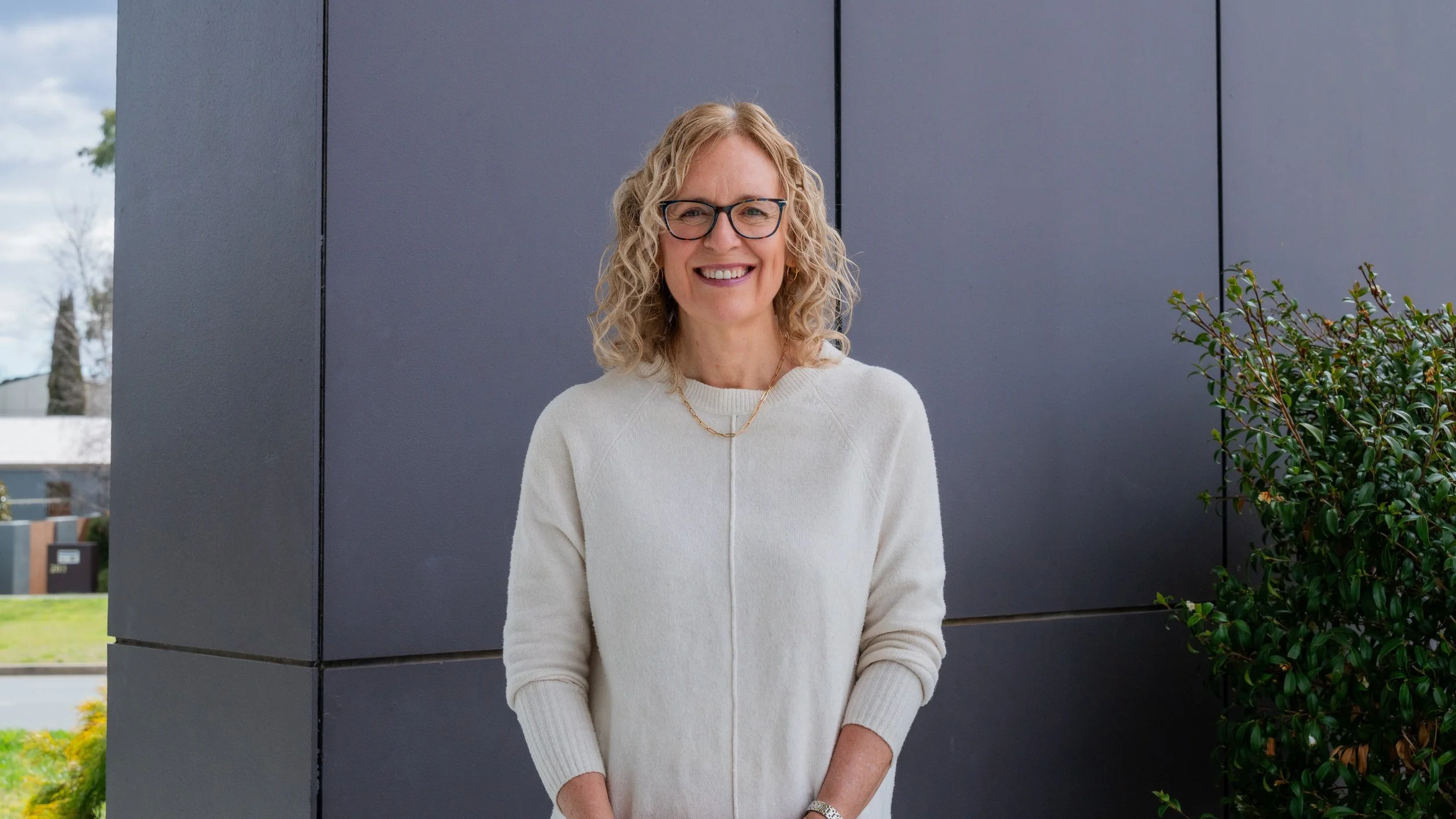 Smiling woman with curly blonde hair, glasses, and a white sweater standing outside against a dark gray wall with greenery on the side.