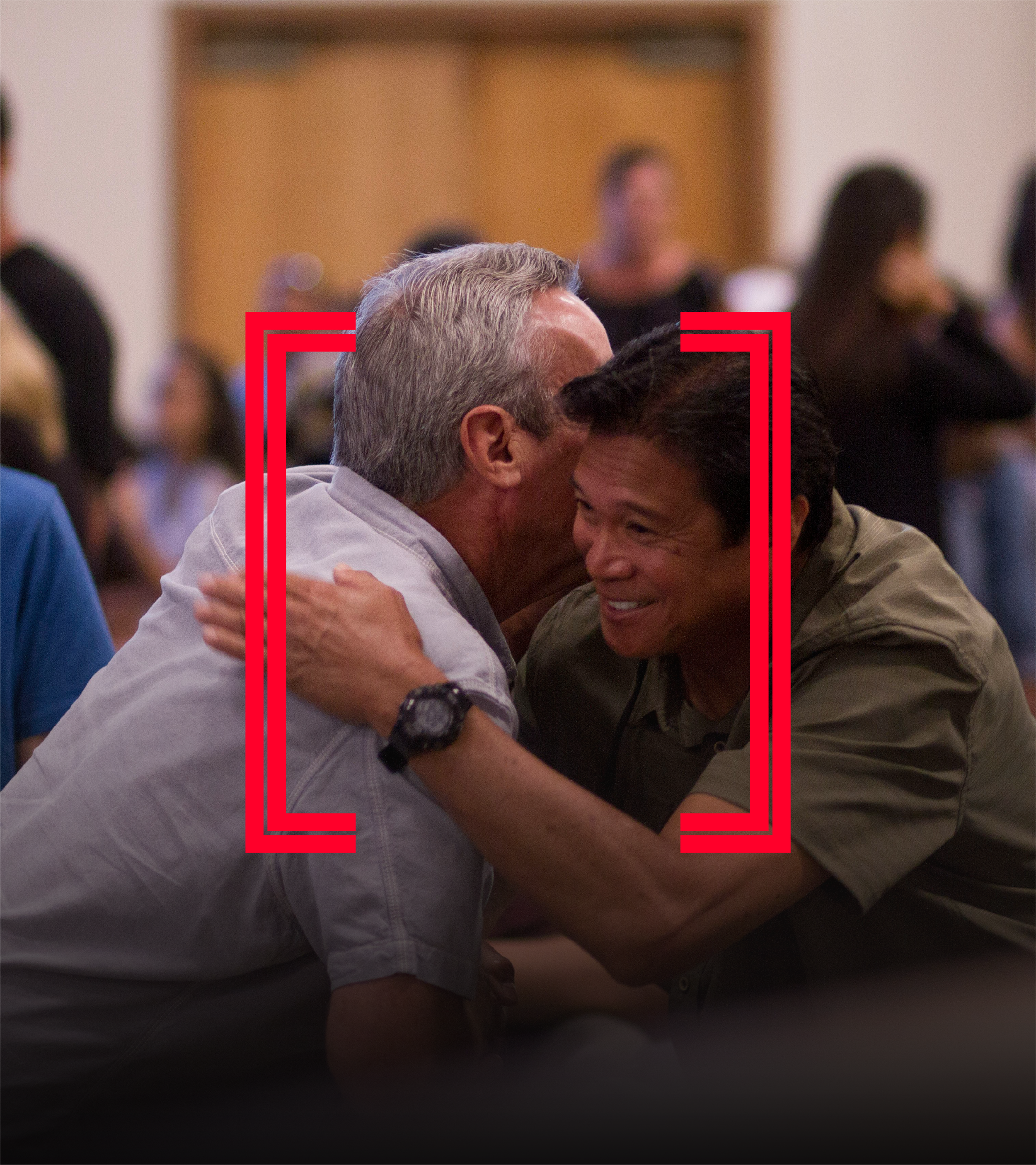 Two men are hugging each other warmly, smiling, in a crowded room. The man on the left has gray hair, while the man on the right has dark hair and glasses. People are seated at tables in the background.