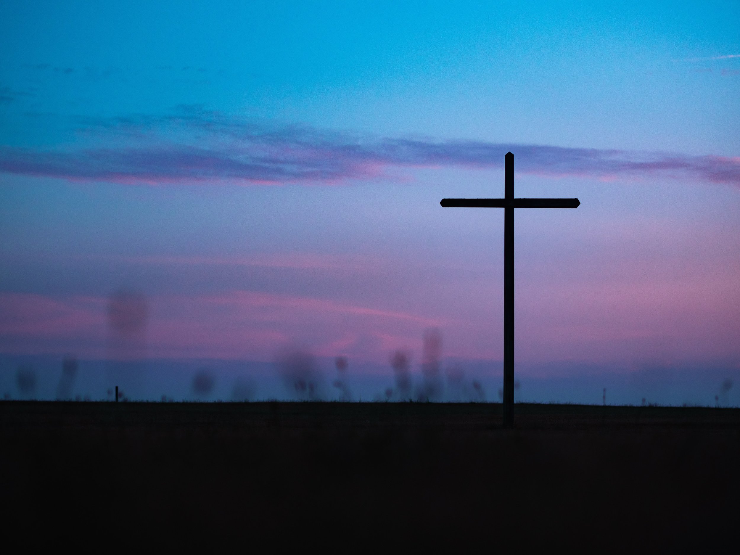 Silhouette of a cross on a flat landscape with a pastel-colored sky and clouds during dusk or dawn.