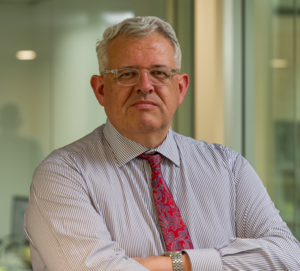 Professional man with glasses, gray hair, wearing a striped shirt and red patterned tie, standing with arms crossed in an office setting.