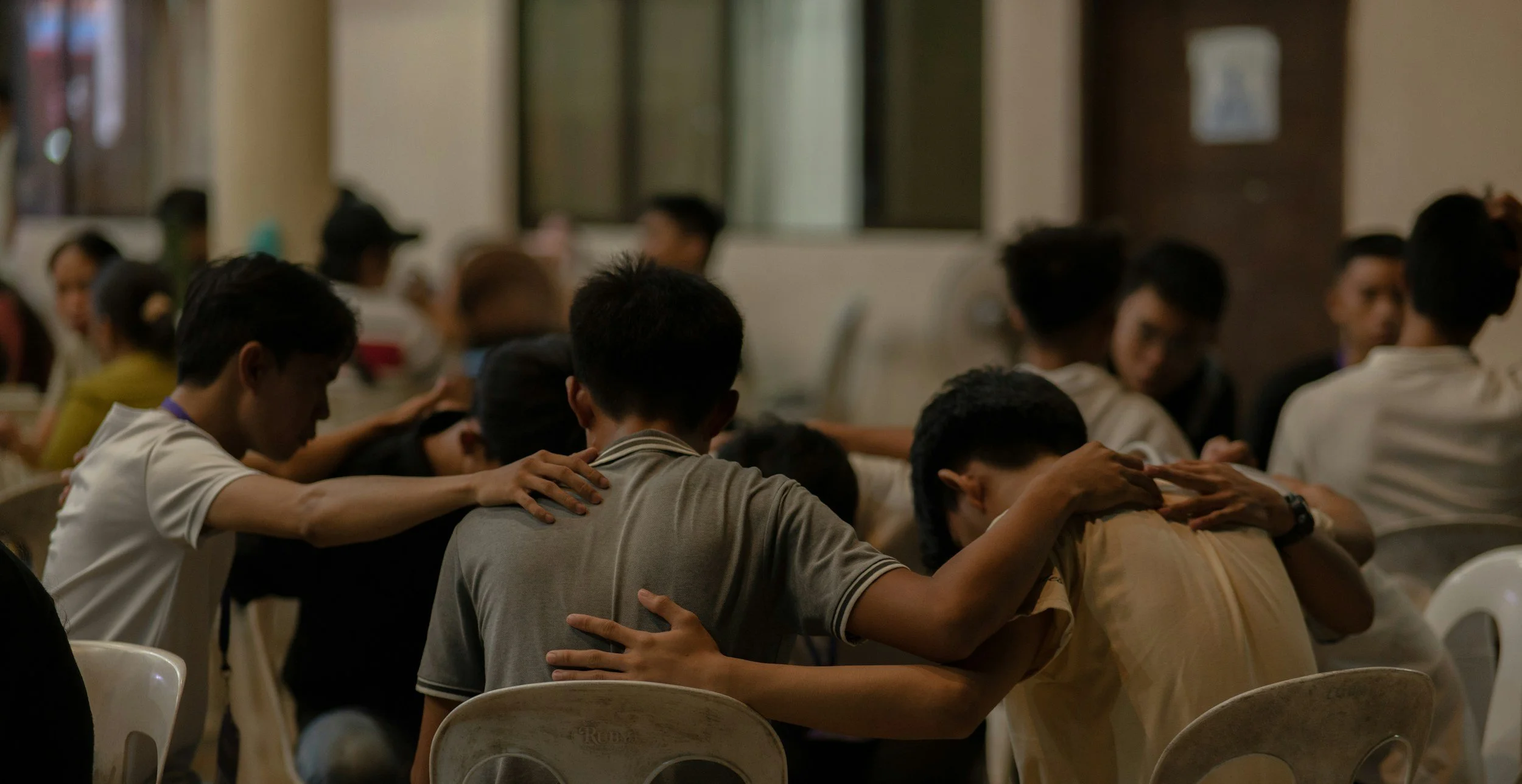 Group of people seated in rows with most having their heads bowed and hands resting on each other's shoulders in a prayer or moment of silence in a dimly lit indoor space.