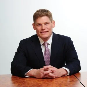 Portrait of a man in a suit sitting at a wooden table against a plain white background.