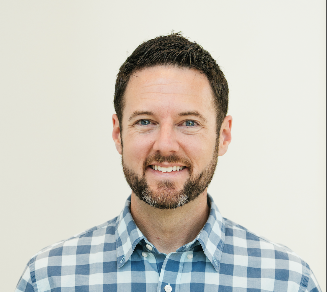 Portrait of a smiling man with dark hair and beard, wearing a blue and white checkered shirt, against a plain light background.