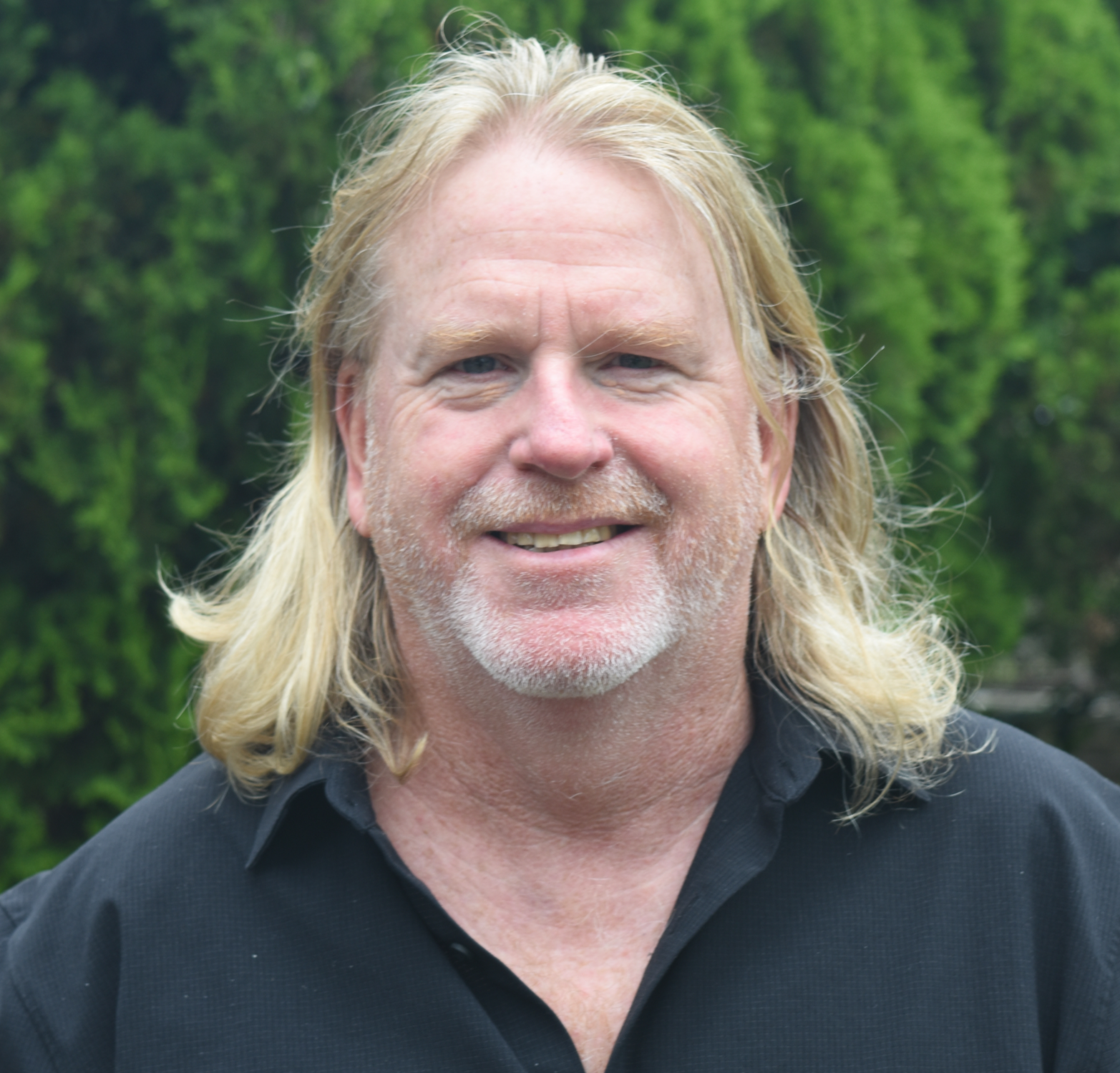 Close-up of a smiling man with long blond hair and a beard, wearing a black shirt, outdoors with green trees in the background.