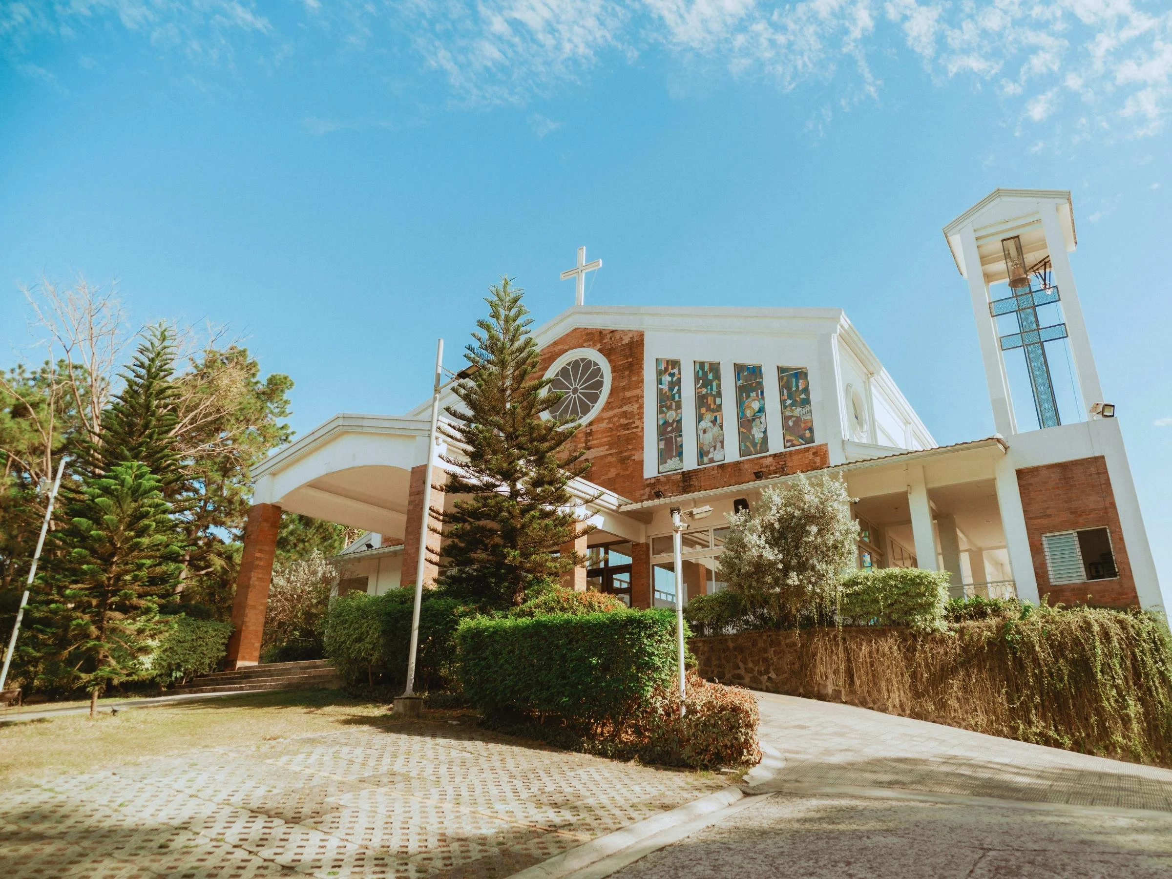 A church building with a white cross on the roof, stained glass windows, and surrounded by trees and shrubbery under a clear blue sky.