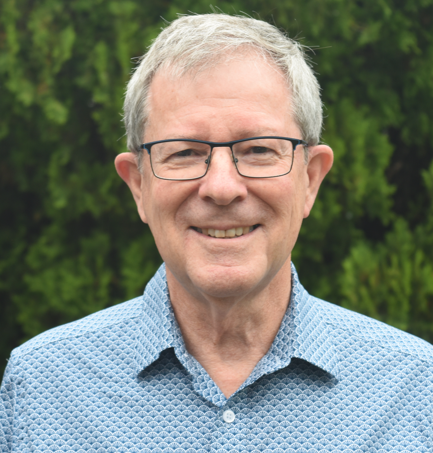 A smiling middle-aged man wearing glasses and a patterned blue shirt outdoors in front of green foliage.