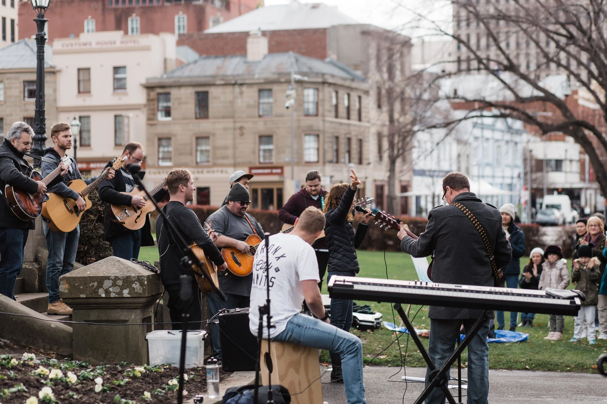A group of musicians performing outdoors in a park with an audience watching. The performers are playing guitars and keyboards, surrounded by trees and buildings in the background.