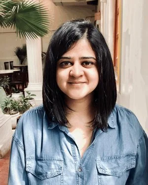 A woman with shoulder-length black hair, smiling, wearing a blue shirt, standing indoors near greenery and wooden architectural elements.