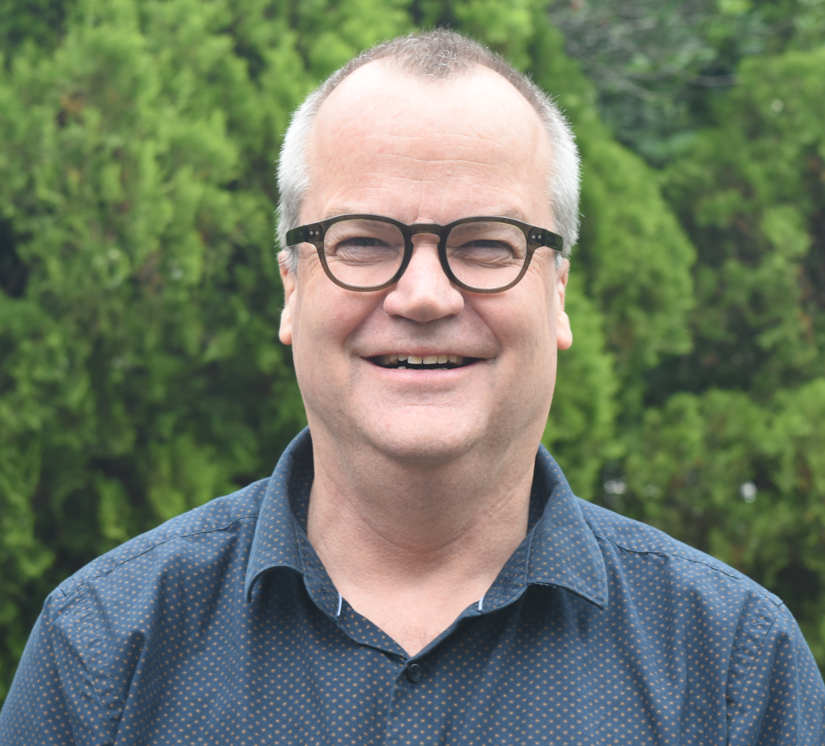 Man wearing glasses smiling outdoors with green trees in the background