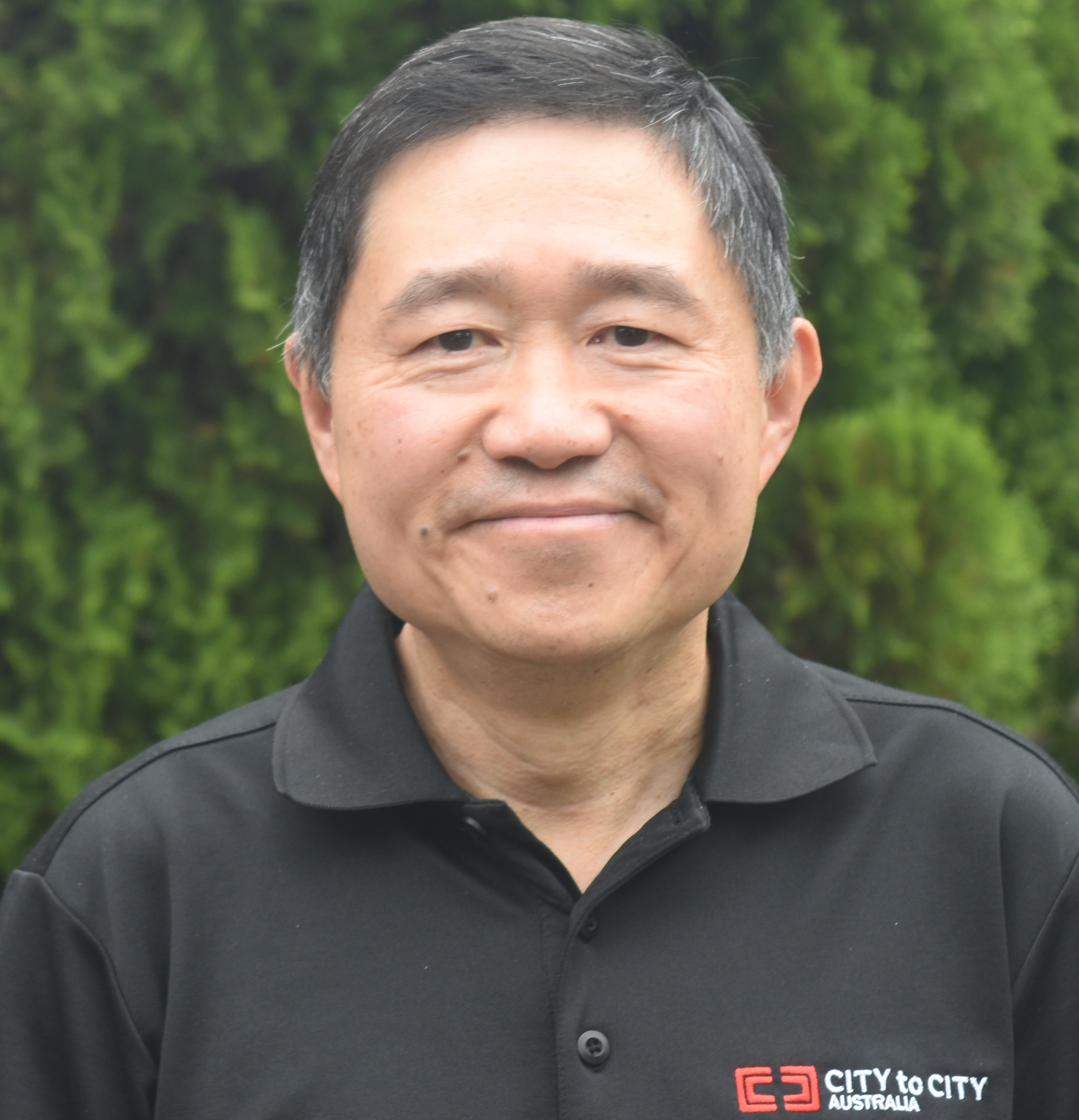 A middle-aged Asian man with short black hair and a slight smile, wearing a black polo shirt with a logo that reads 'City to City Australia,' standing outdoors with a blurry green foliage background.
