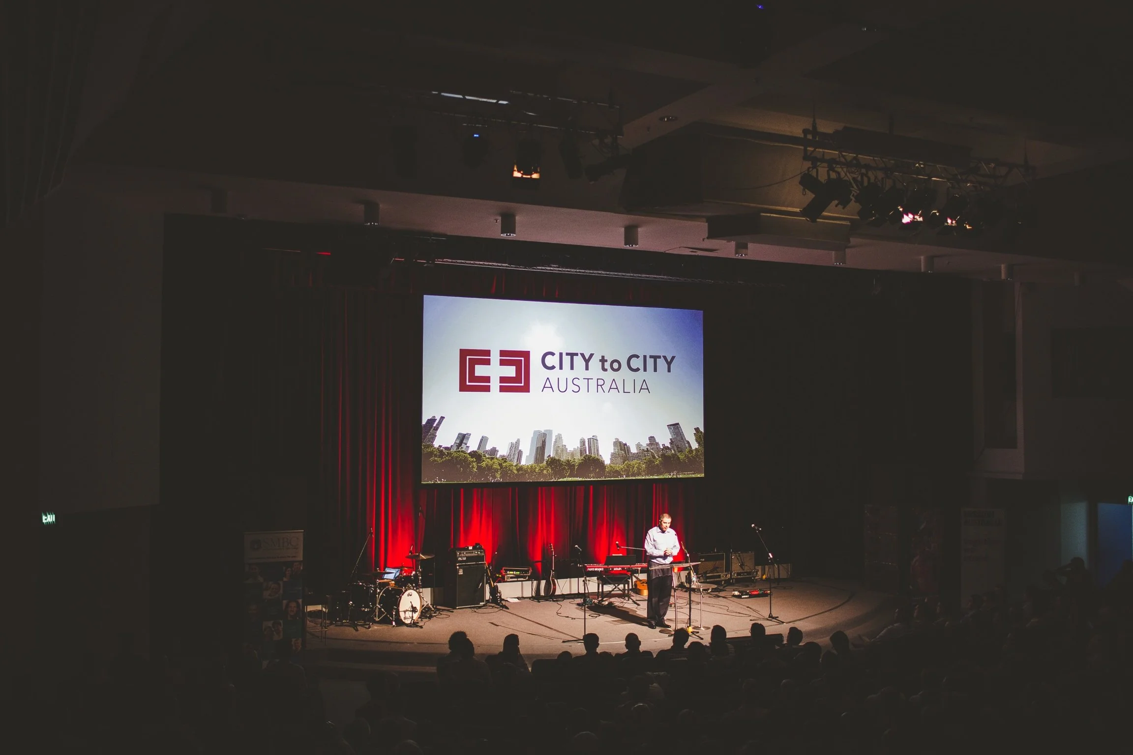 An indoor stage with a large screen displaying the logo 'CITY to CITY AUSTRALIA' and a cityscape background. There are musical instruments and a person speaking at a microphone, with an audience seated in front.