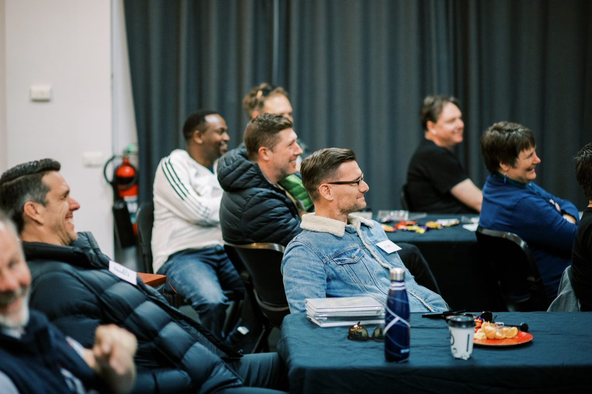 A group of people sitting at tables, smiling and laughing during a meeting or seminar. There are notebooks, a water bottle, and snacks on the table.