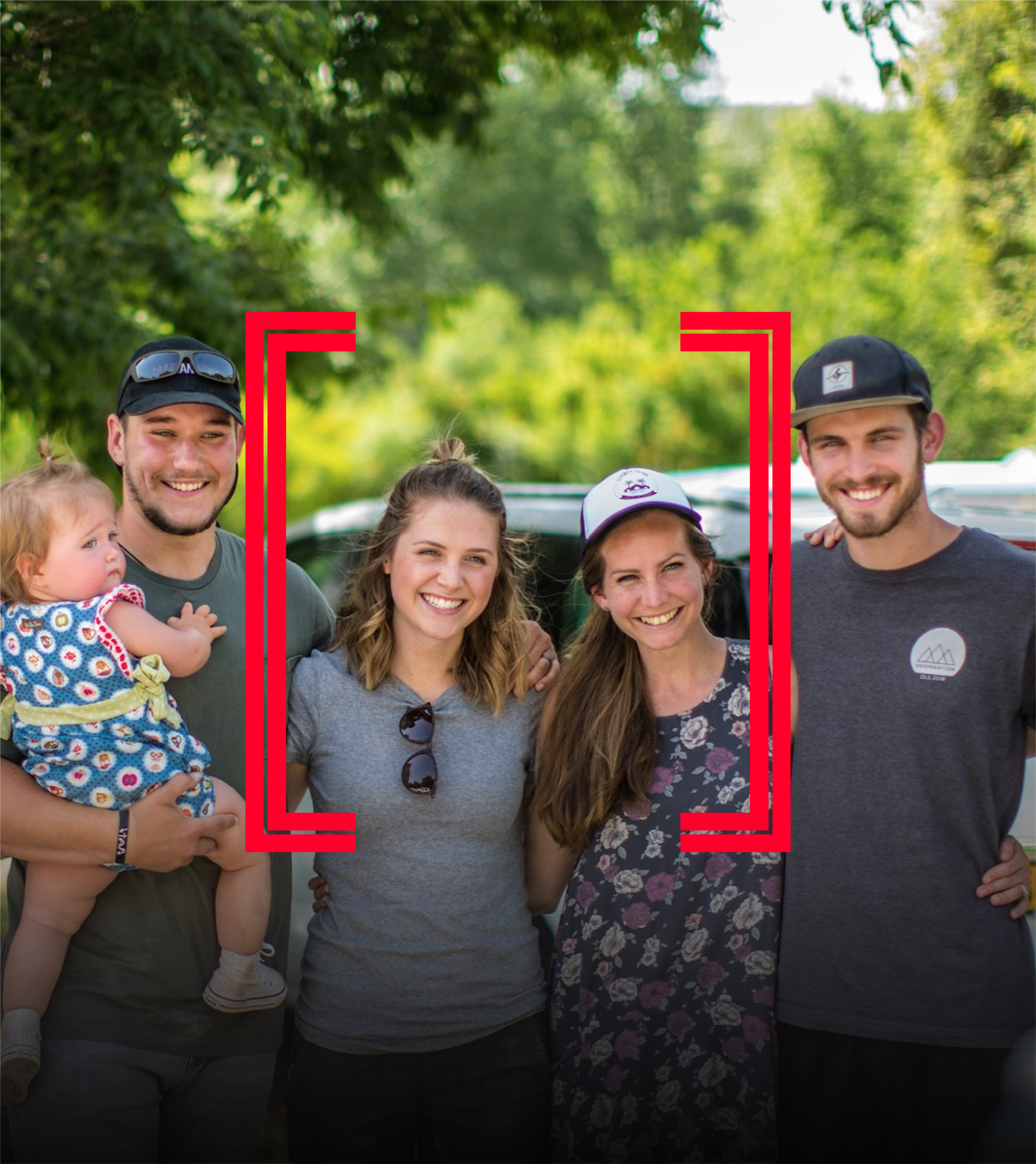 A group of smiling people outdoors, including men, women, and a young girl, with green trees in the background, some wearing hats and sunglasses.