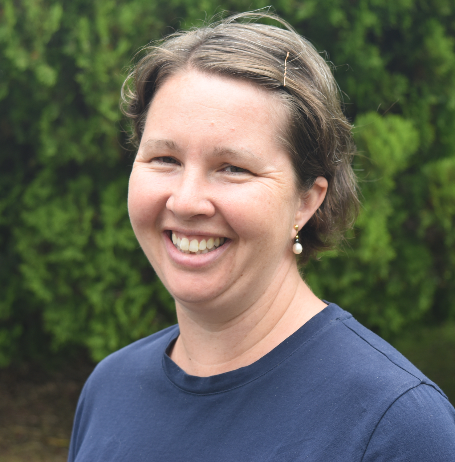 A smiling woman with short brown hair, wearing pearl earrings and a navy blue shirt, standing outdoors with greenery in the background.