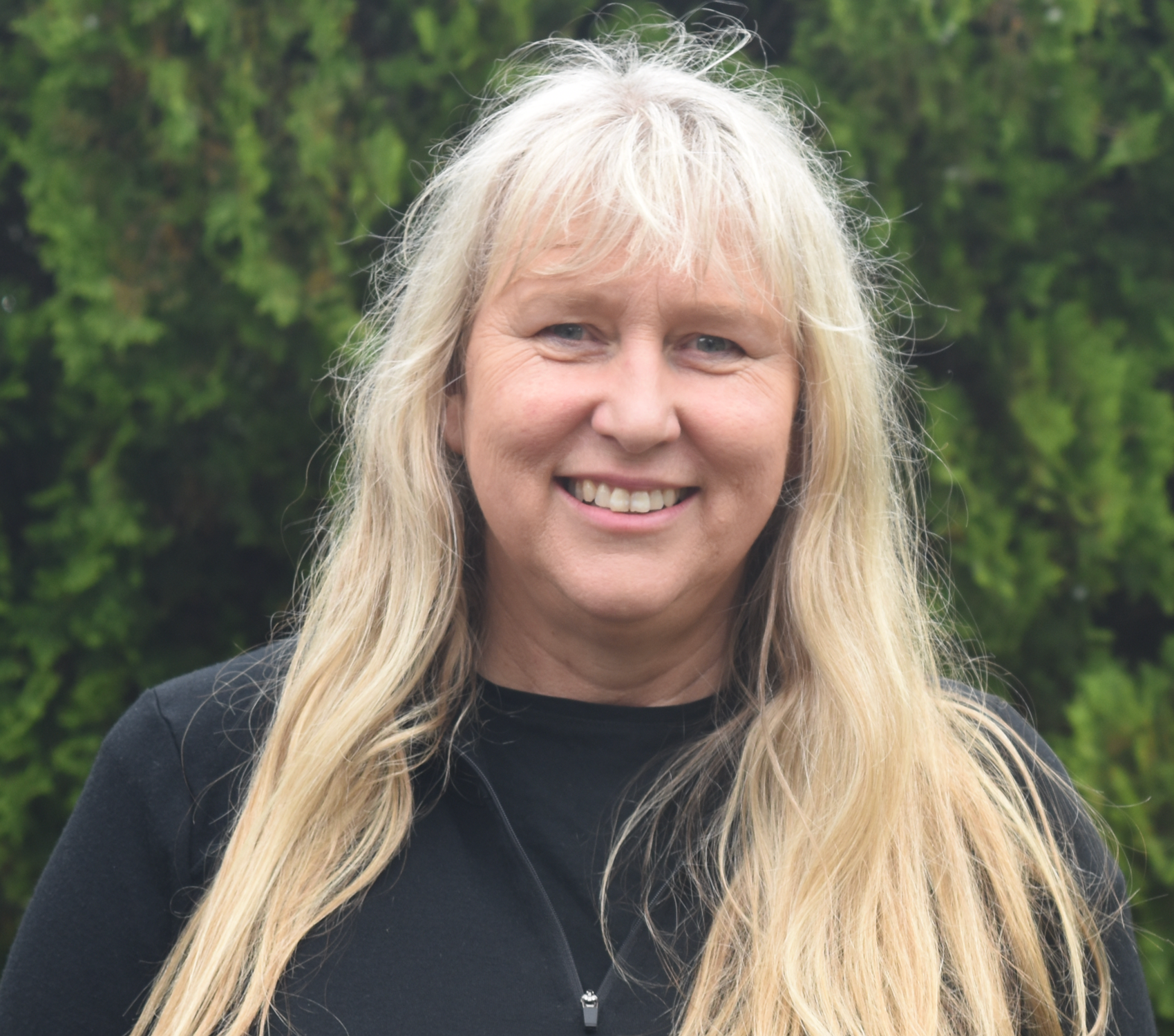 A woman with long blonde hair, smiling, wearing a black top, outdoors with green foliage in the background.