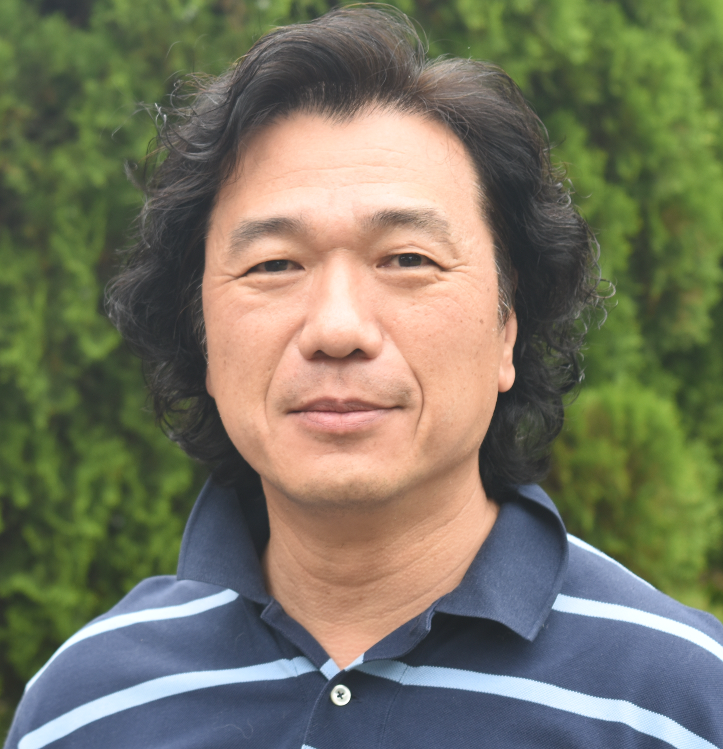 A man with curly black hair, wearing a navy blue polo shirt with white stripes, standing outdoors with greenery in the background.