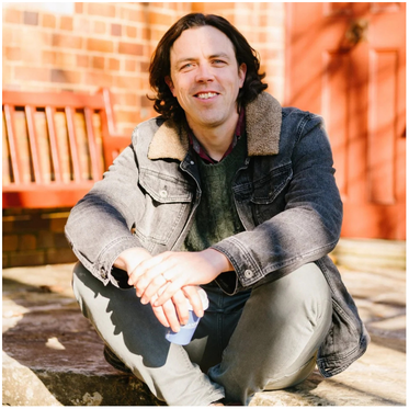 A man with shoulder-length dark hair, wearing a denim jacket with a shearling collar, sitting outdoors on a brick patio with a wooden bench in the background.