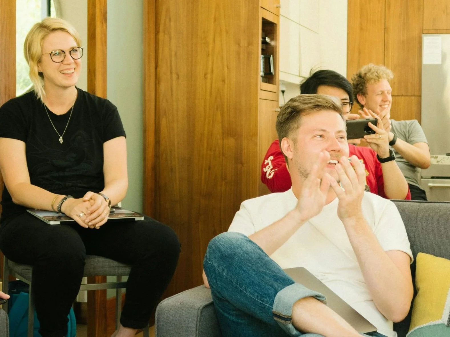 Group of four young adults, two men and two women, enjoying a social gathering indoors. They are sitting on a couch and a chair, laughing and smiling, with one man holding a smartphone, capturing a moment.