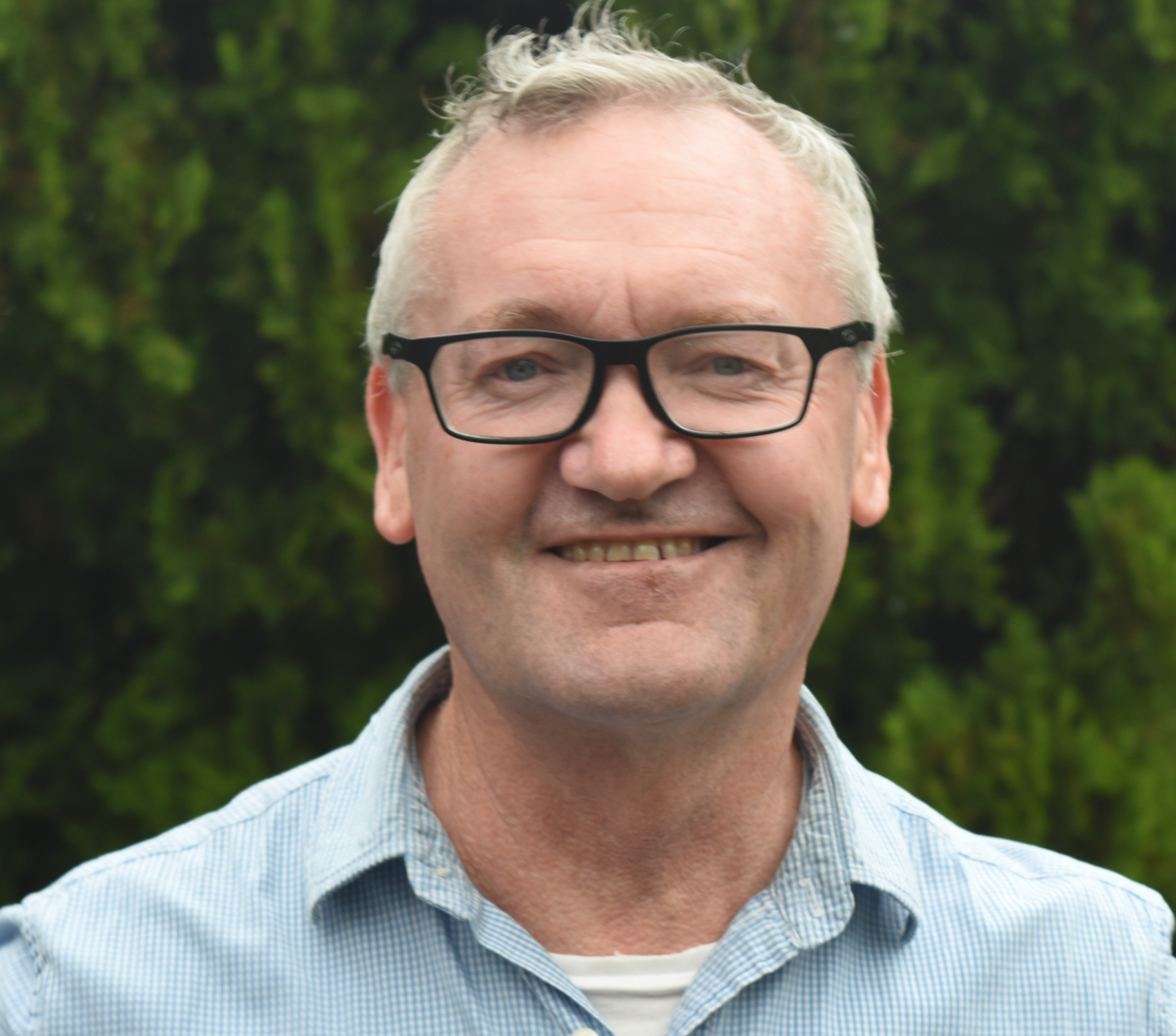 Close-up of a smiling middle-aged man with gray hair and glasses, wearing a blue checkered shirt, outdoors with green foliage in the background.