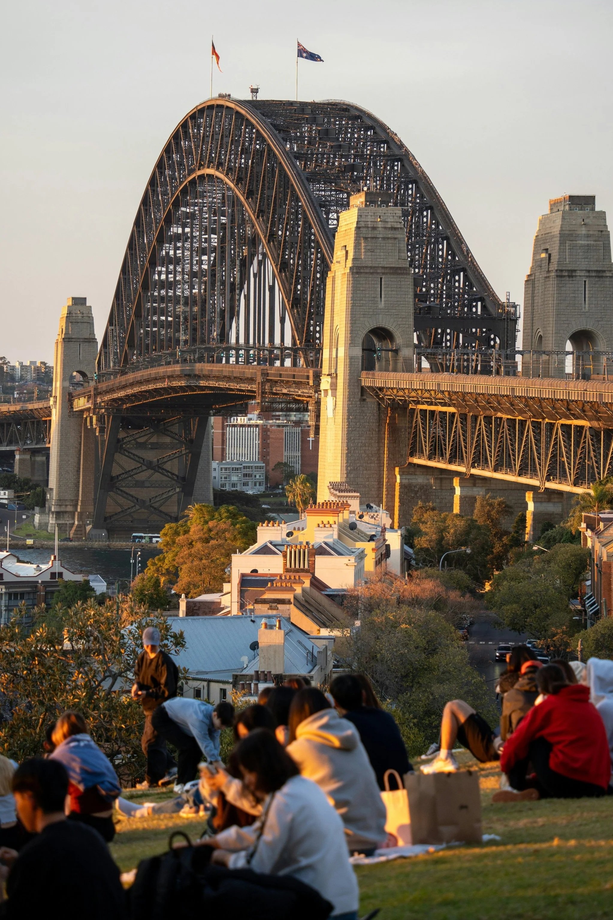 A group of people sitting on the grass in front of the Sydney Harbour Bridge, with some looking at their phones and others talking, during sunset.