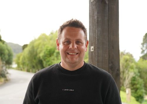 A man smiling outdoors next to a wooden post, with trees and a cloudy sky in the background.