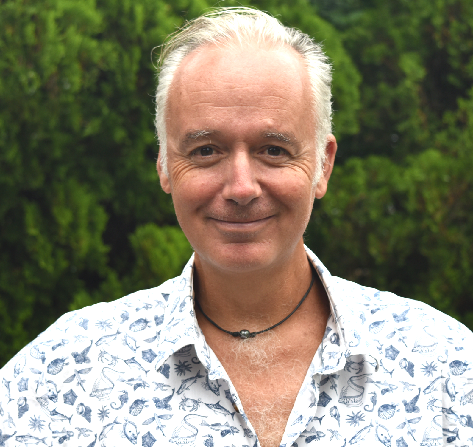 A middle-aged man with short gray hair smiling outdoors in front of green trees, wearing a white patterned shirt and a black necklace.