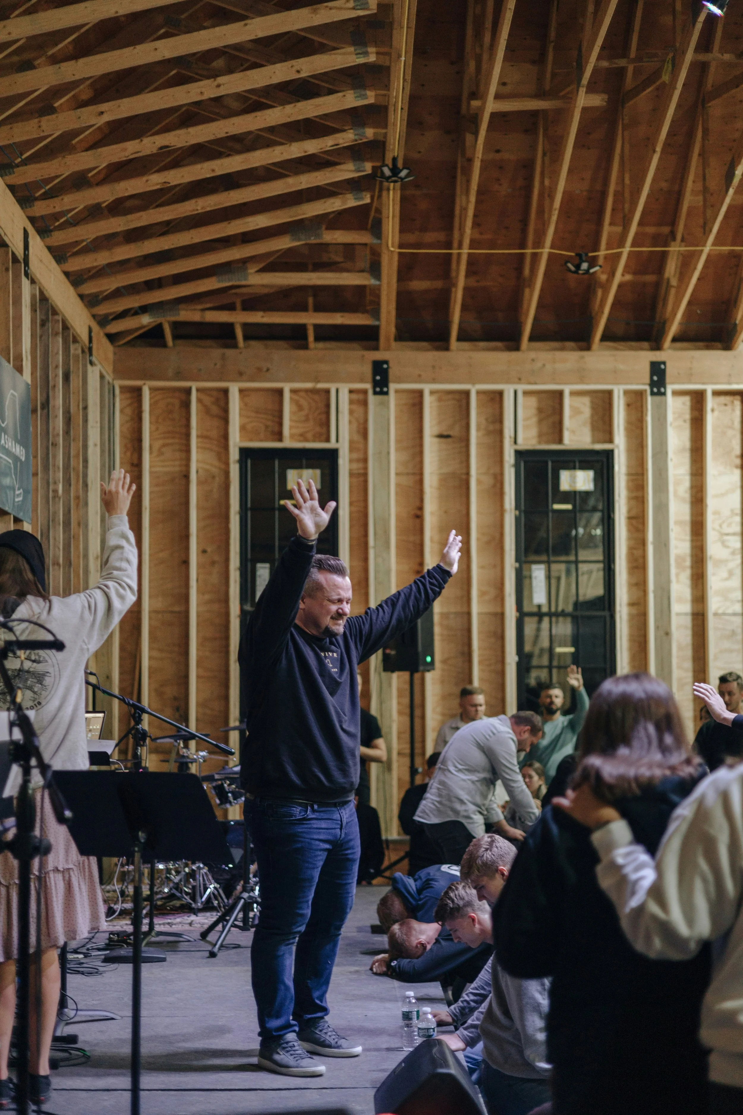 People in a church or community center praying, some standing and some kneeling on the floor, with a man raising his hands in worship, wooden walls, and windows in the background.