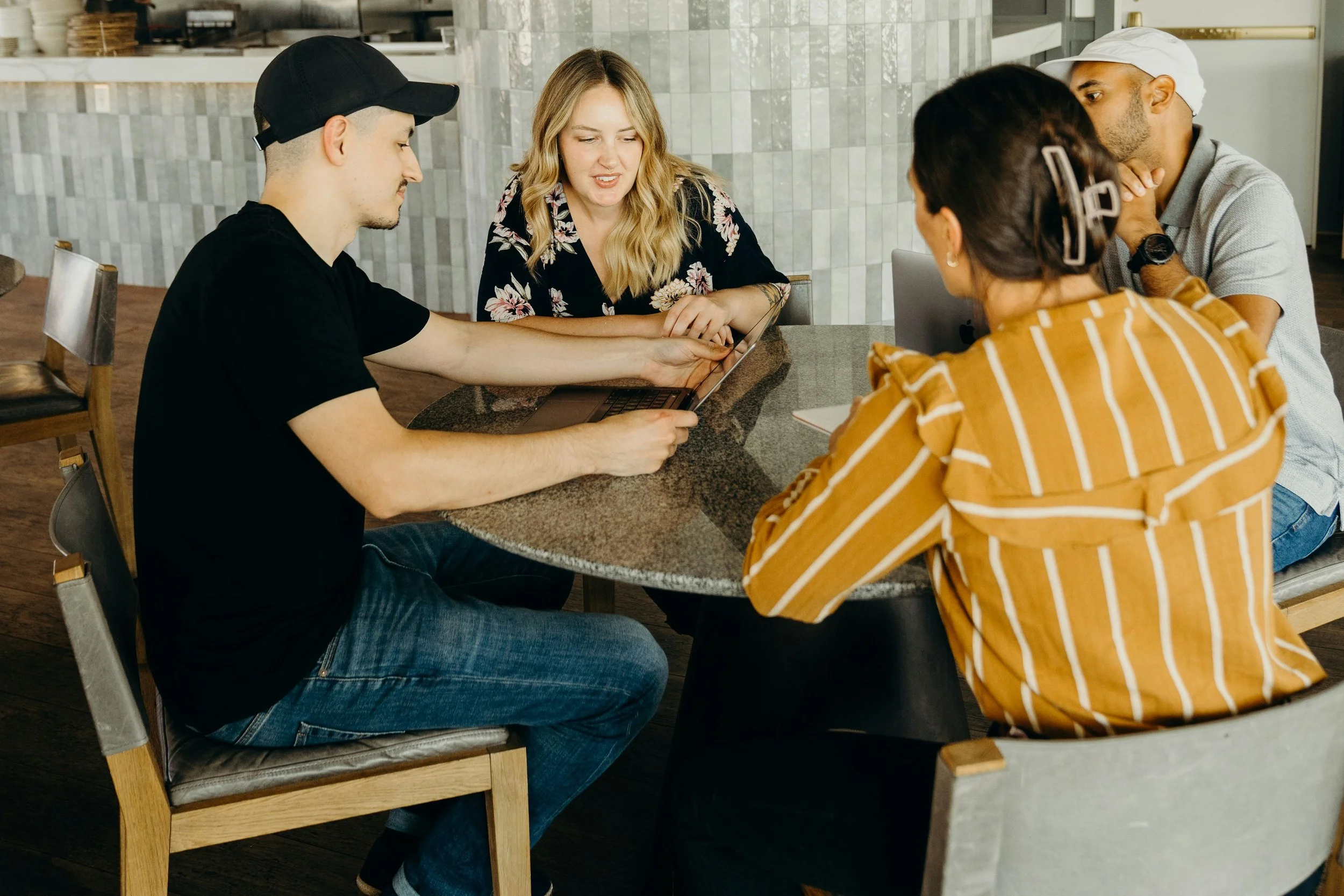 A group of five people sitting at a restaurant table looking at a laptop screen, engaged in conversation.