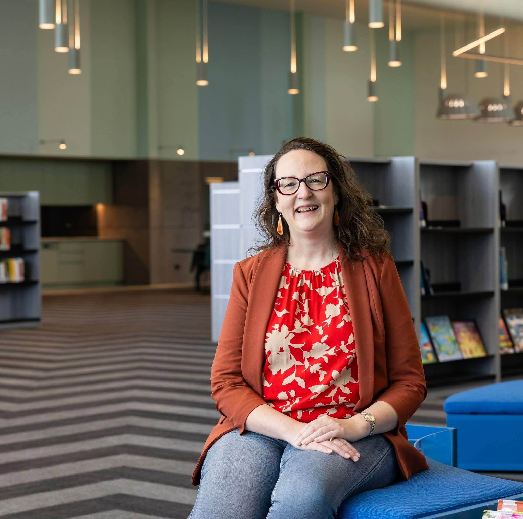 A smiling woman with glasses sitting on a blue bench in a modern library or office with green walls and hanging lights.