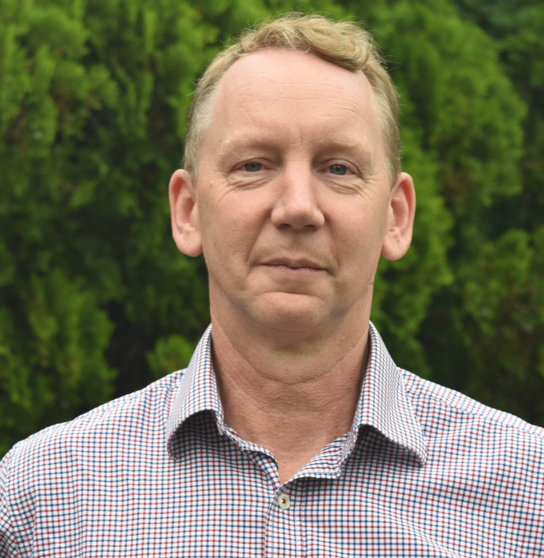 Close-up of a middle-aged man with short, light-colored hair, wearing a checkered shirt, standing outdoors with green trees in the background.