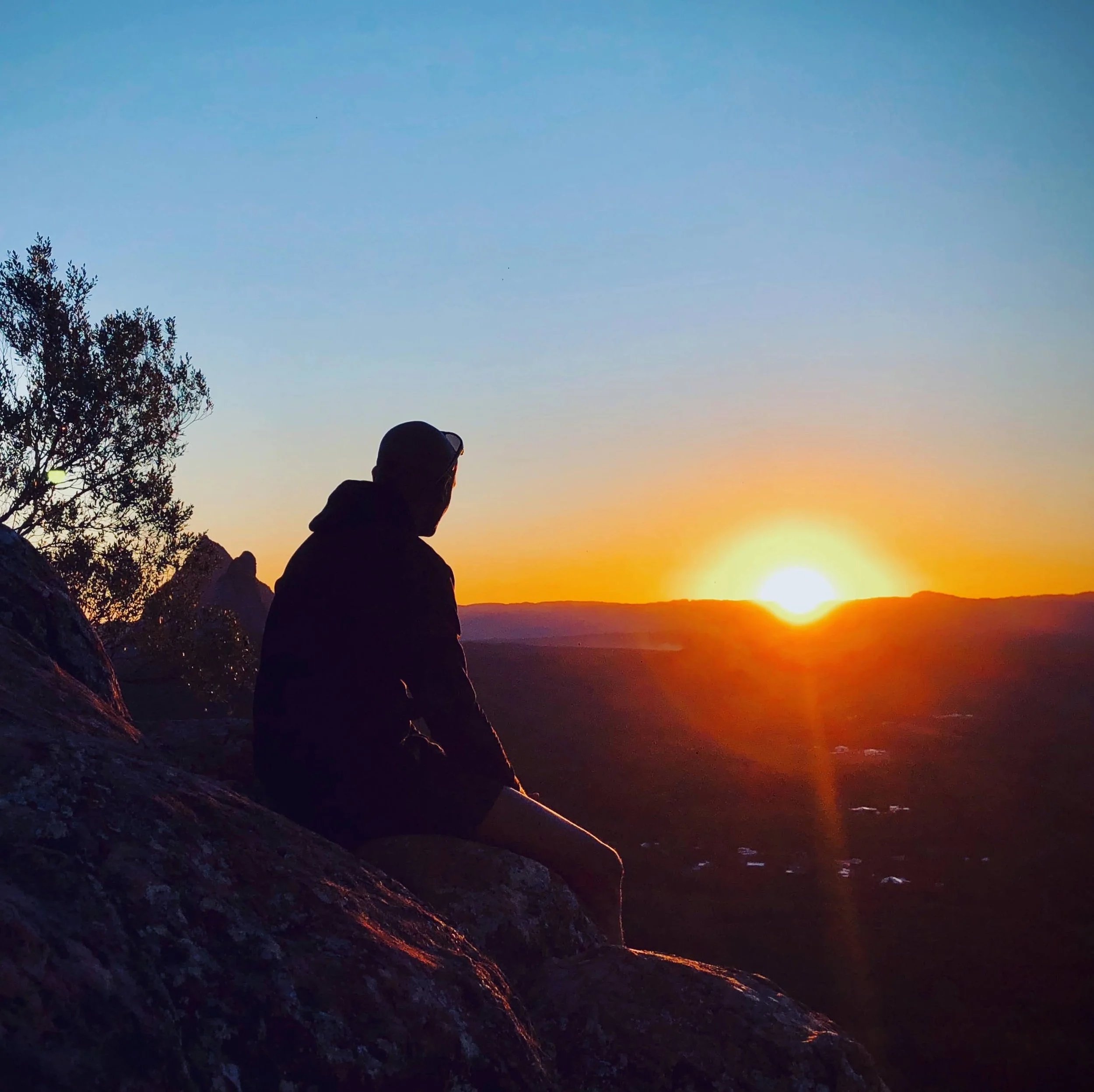 Silhouettes of two people sitting on rocks, watching a sunset over a mountainous landscape with a tree on the left side of the image.