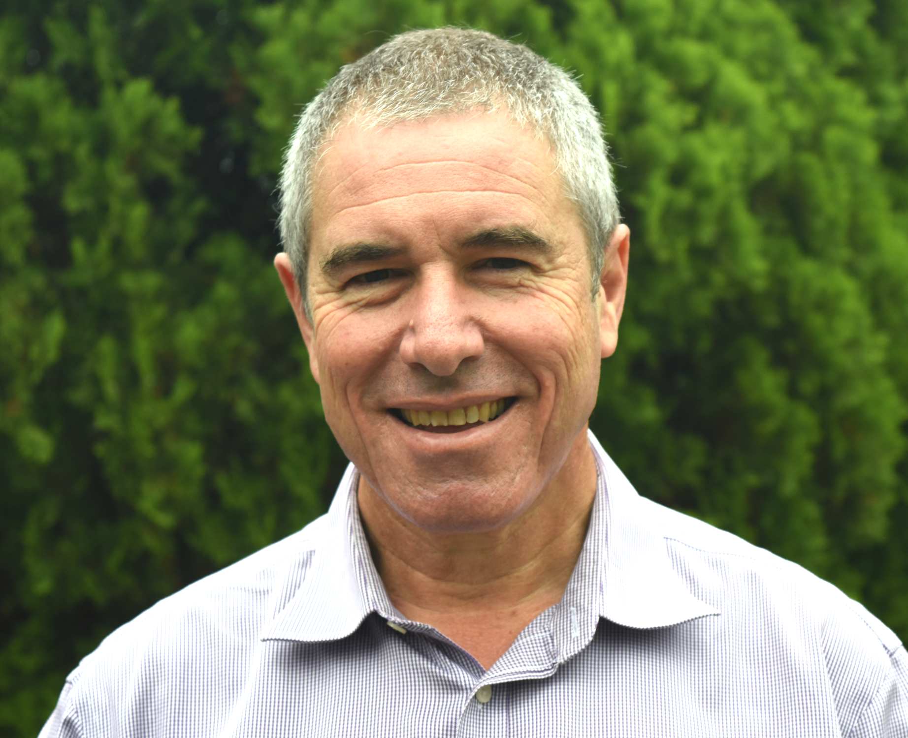 Close-up of a smiling middle-aged man with short gray hair, wearing a light-colored striped shirt, standing outdoors with green trees in the background.