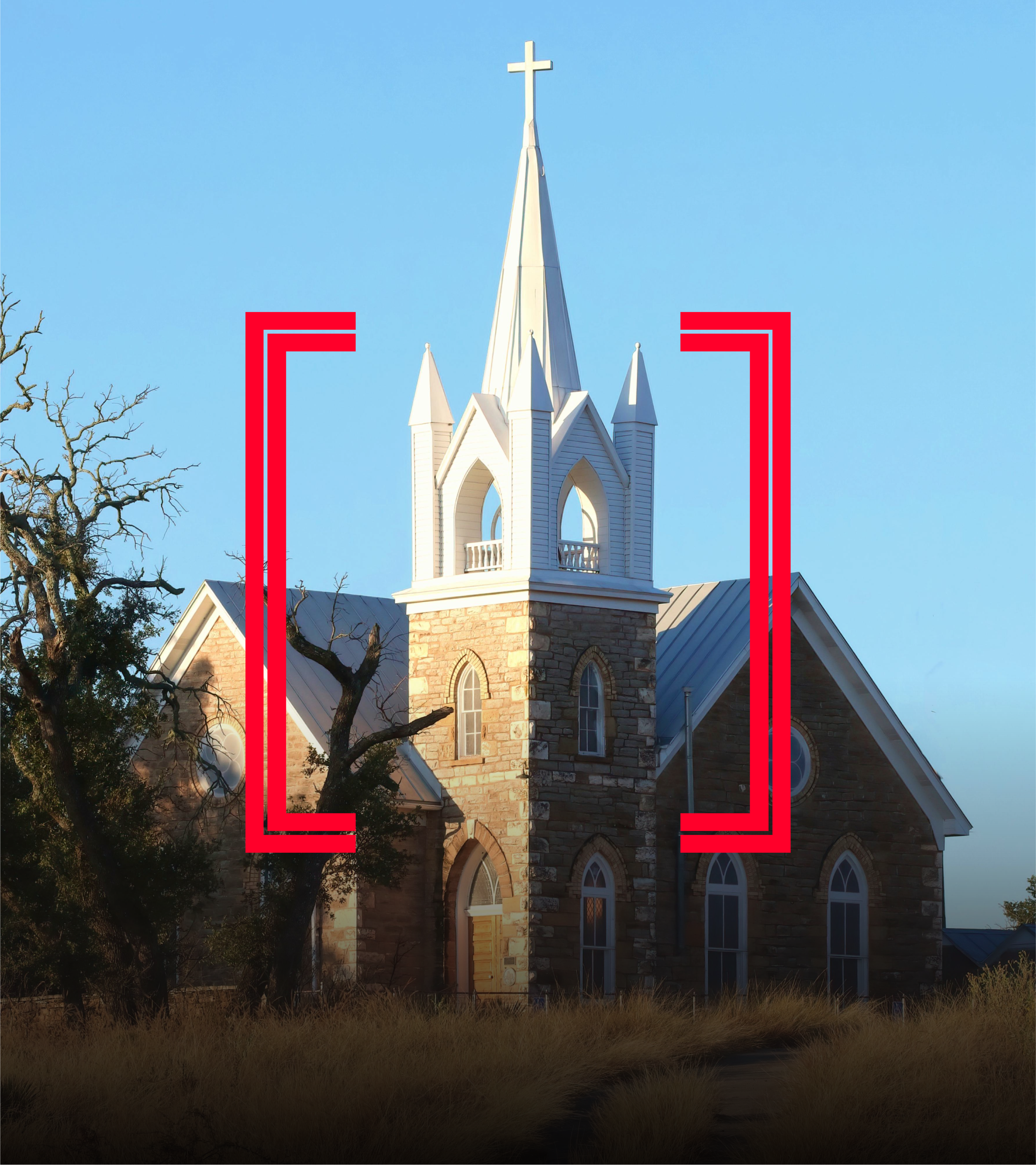 A white church with a tall steeple topped with a cross, brick and stone walls, pointed arch windows, and a blue sky background, with trees and grass in the foreground.