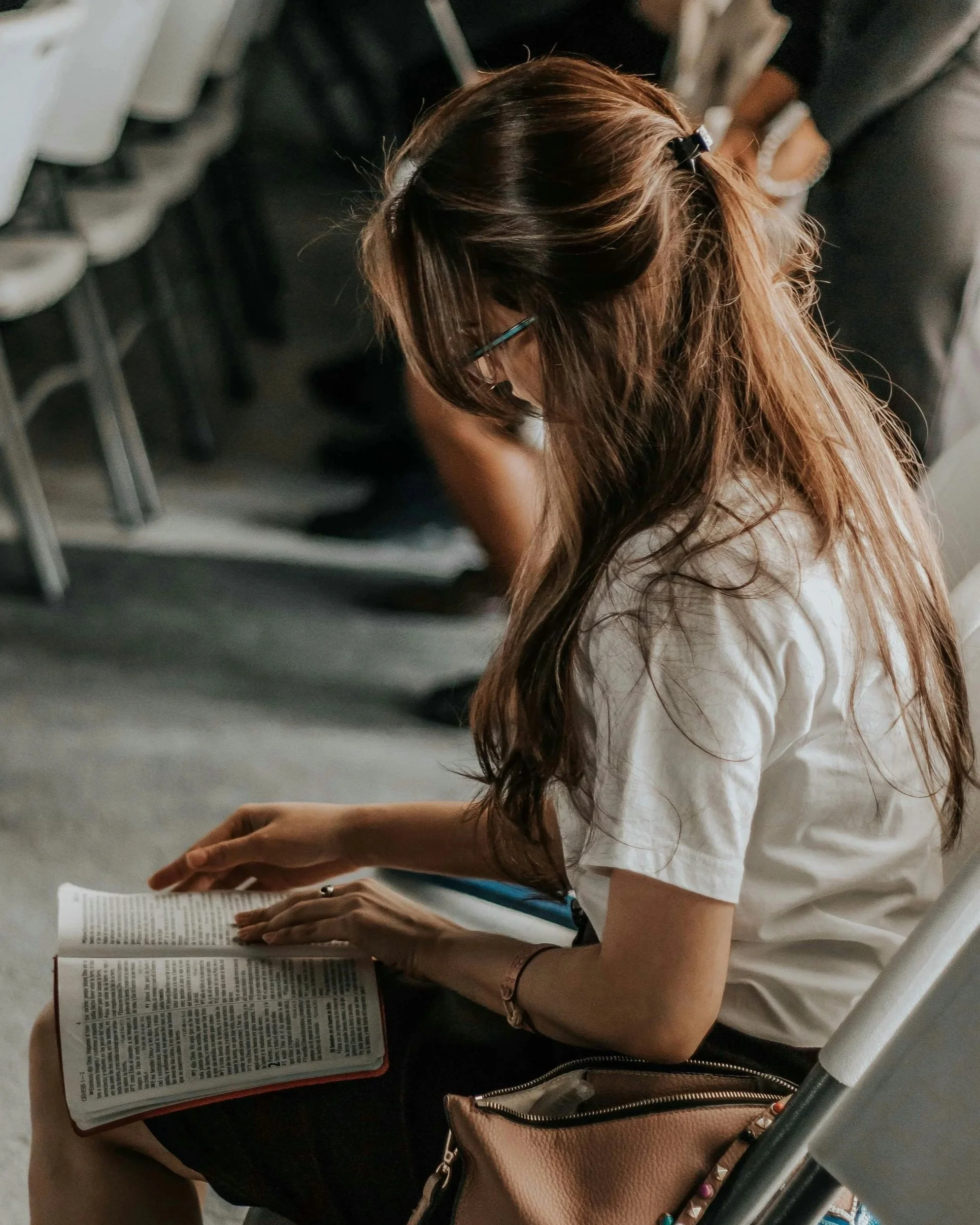 A woman with long brown hair and glasses, wearing a white shirt, is sitting at a table reading a book.