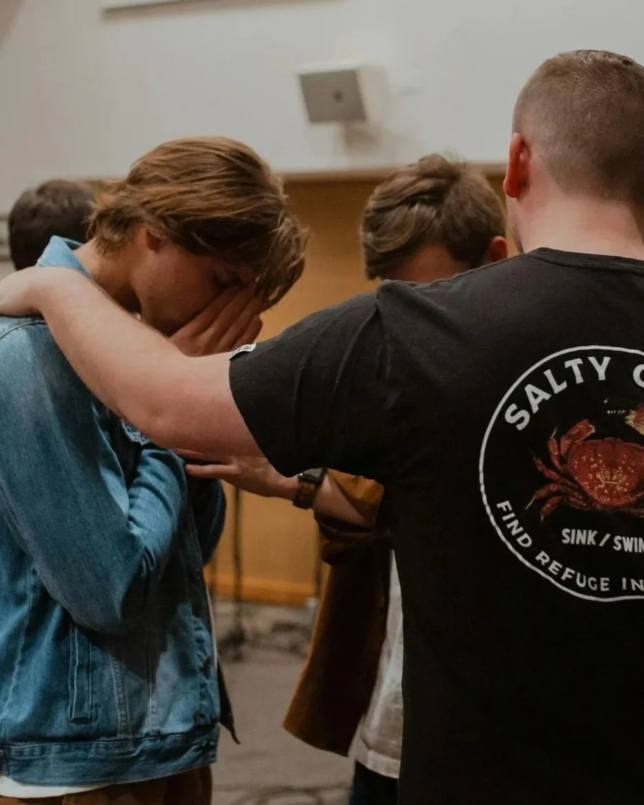 A group of young men participating in a prayer or moment of reflection, with one young man holding his hands over his face, while others have their hands on his shoulders. The setting appears to be indoors, possibly in a church or community center.