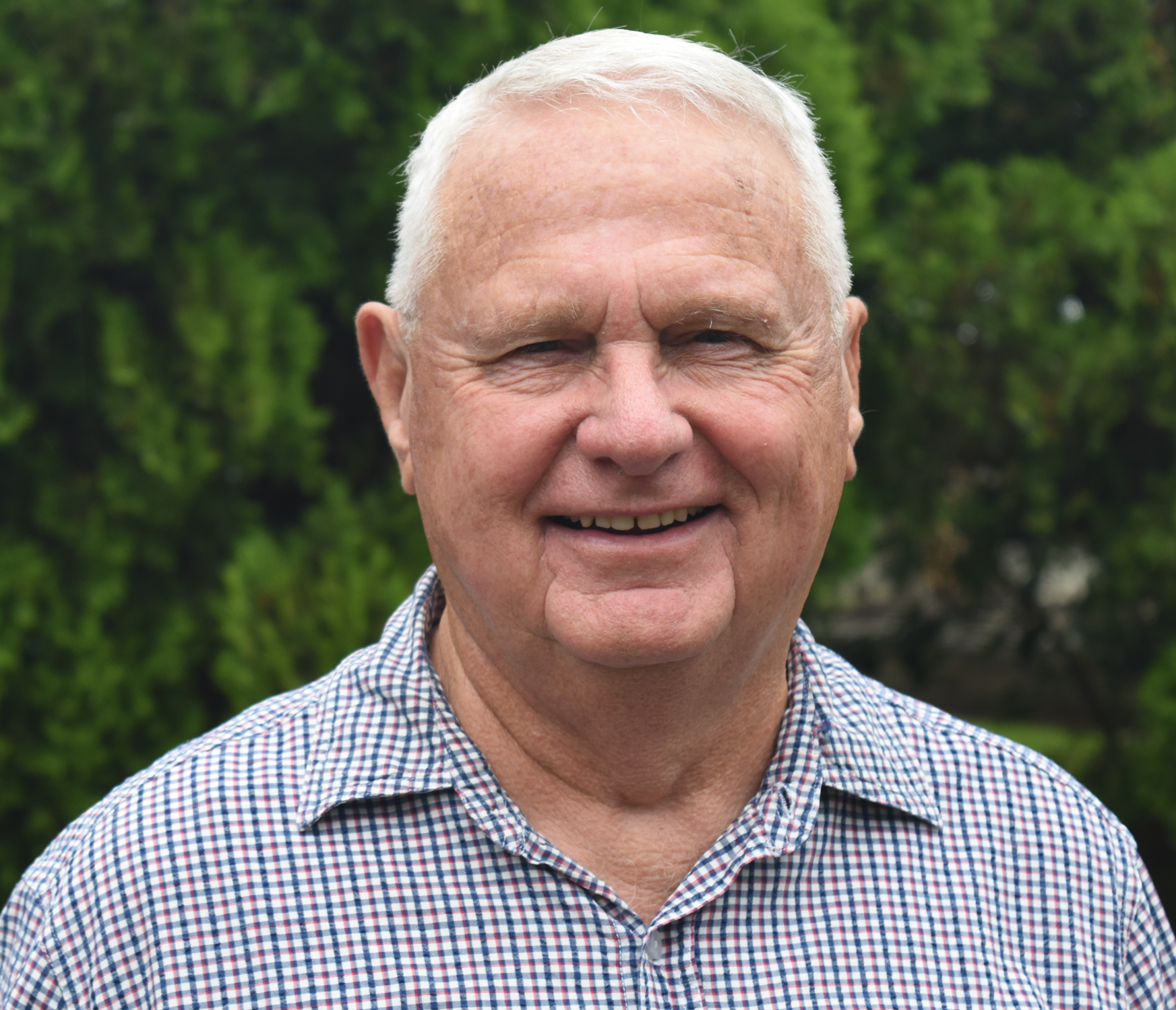 Close-up of an elderly man with white hair, smiling, wearing a checkered shirt, outdoors with green trees in the background.