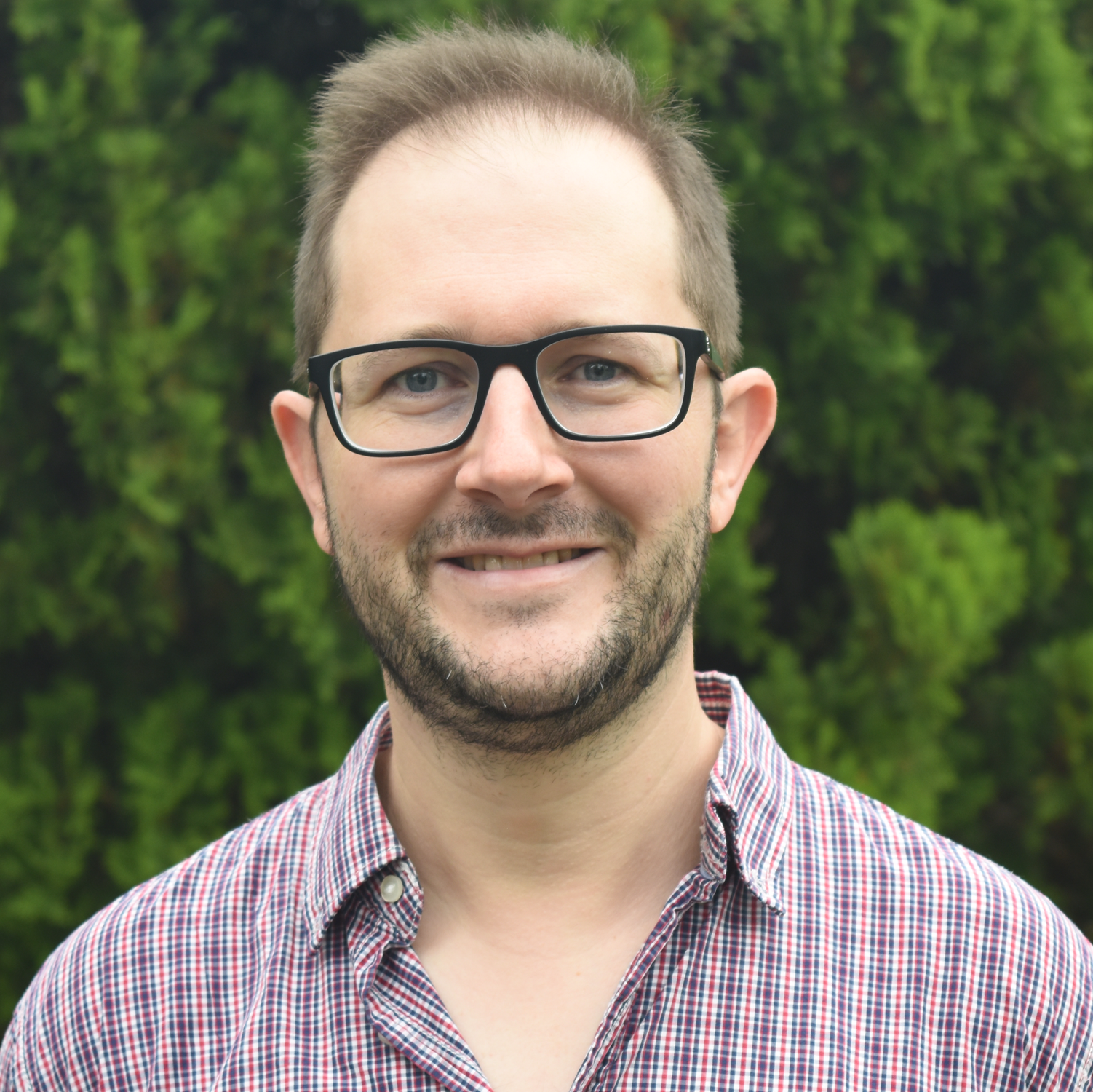 A man with glasses and a short beard smiling outdoors, wearing a checked shirt, with green foliage in the background.