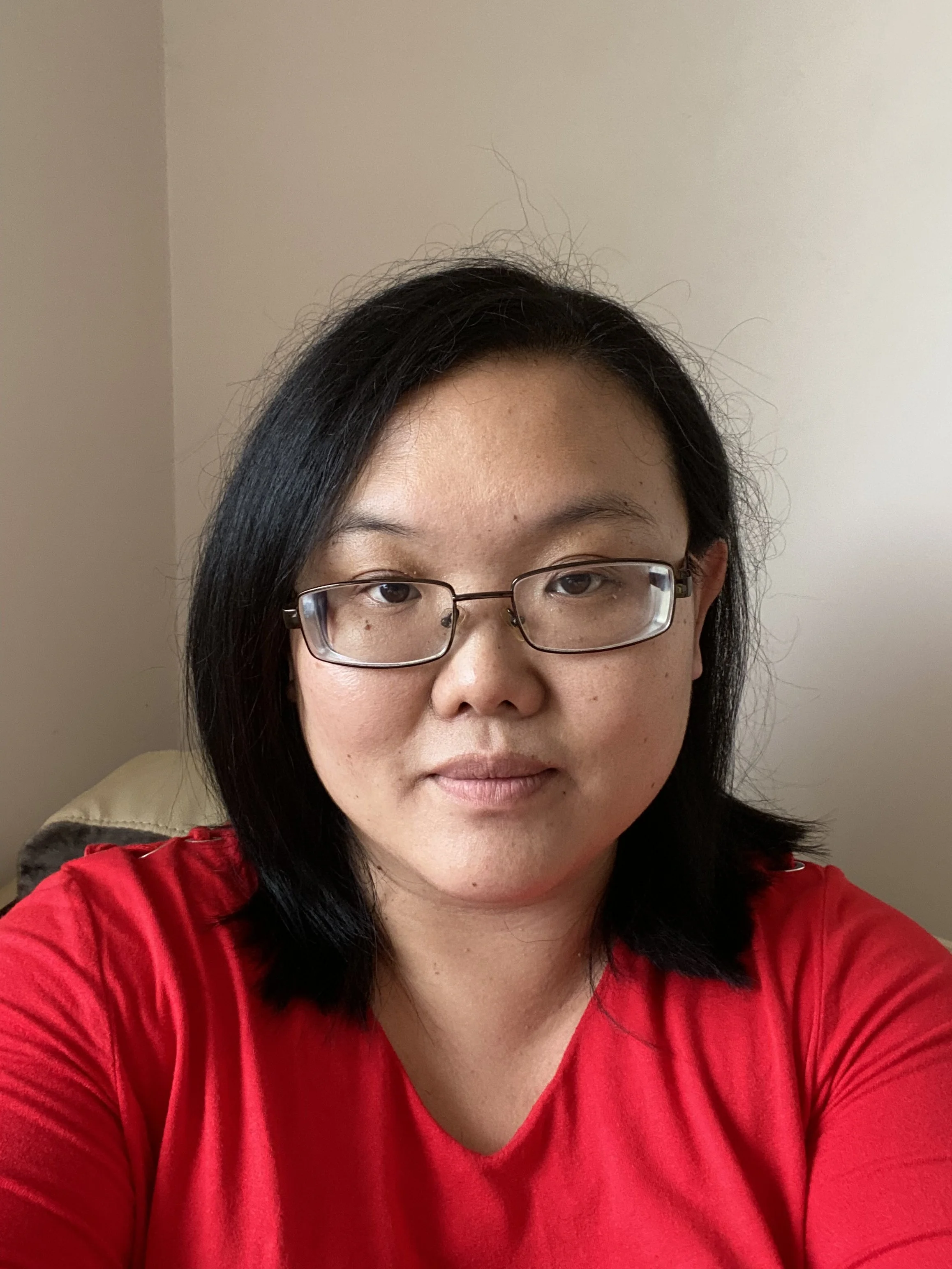 Close-up of a woman with black hair, glasses, and wearing a red top, against a plain beige wall.