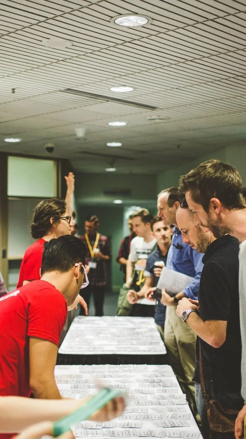 Group of people gathered around tables with papers or badges, engaged in conversation at an indoor event or conference.
