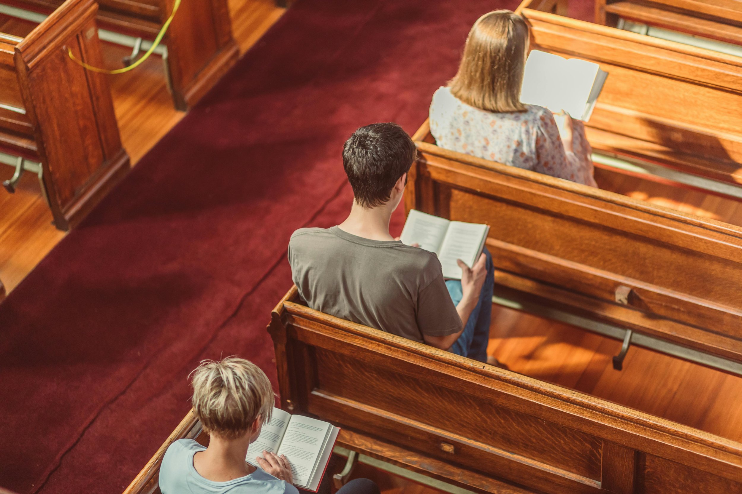 People sitting in pews reading books in a church or chapel with wooden benches and red carpeted aisle.