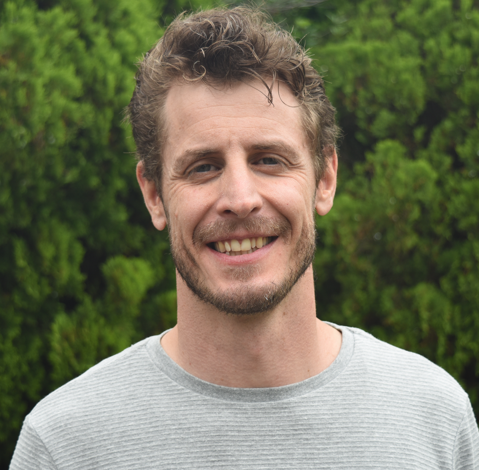 Smiling man with curly light brown hair and beard, wearing a light gray shirt, standing outdoors with green foliage in the background.