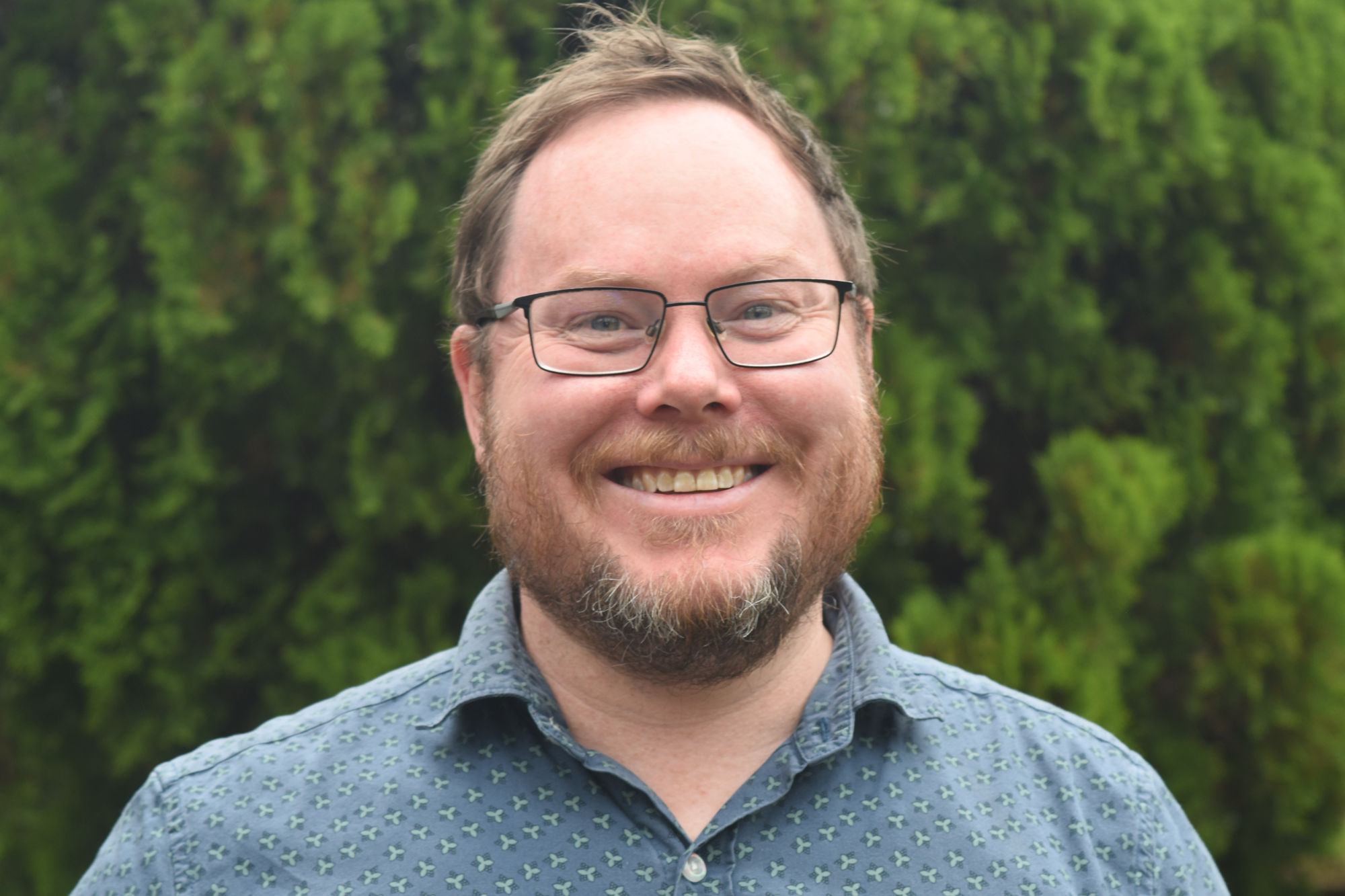 Close-up of a smiling man with glasses and a beard, wearing a patterned button-up shirt, standing outdoors with greenery in the background.