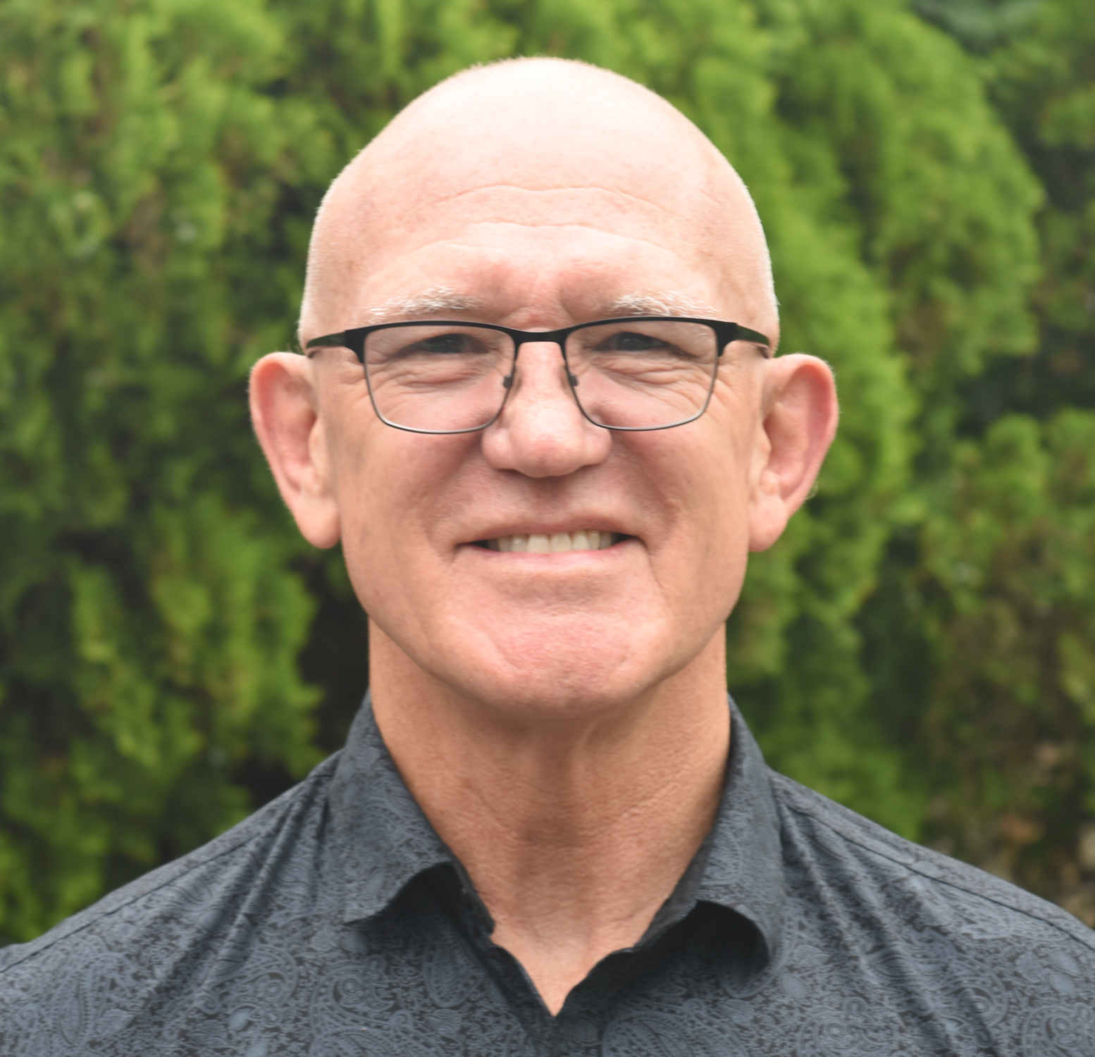 A middle-aged man with a shaved head and glasses smiling outdoors with green trees in the background.