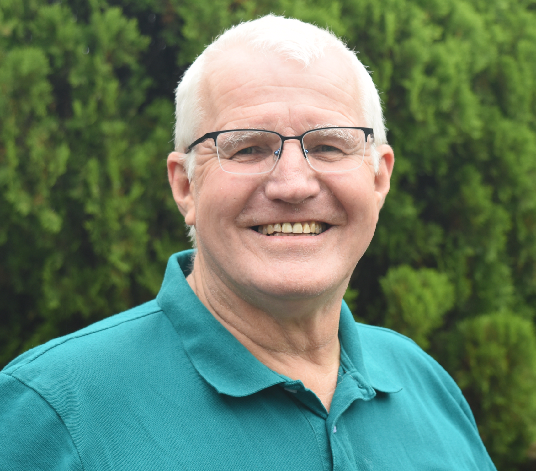 A smiling older man with white hair and glasses, wearing a teal polo shirt, standing outdoors in front of green trees.