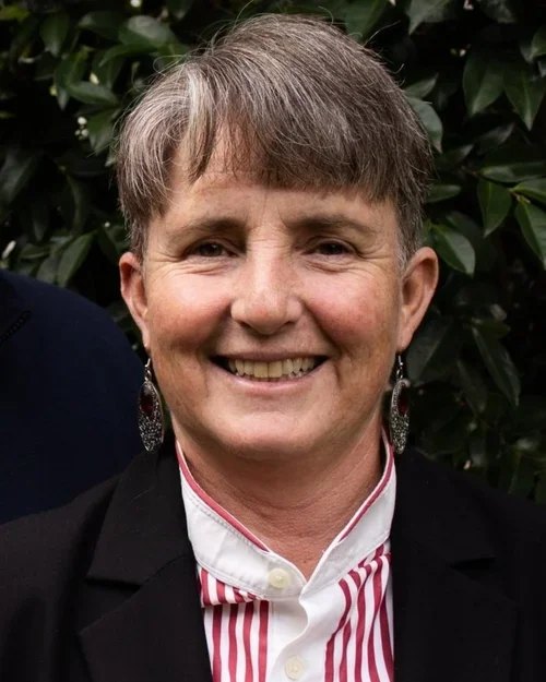 A smiling woman with short gray hair wearing earrings, a black blazer, and a white shirt with red stripes, standing outdoors in front of green bushes.