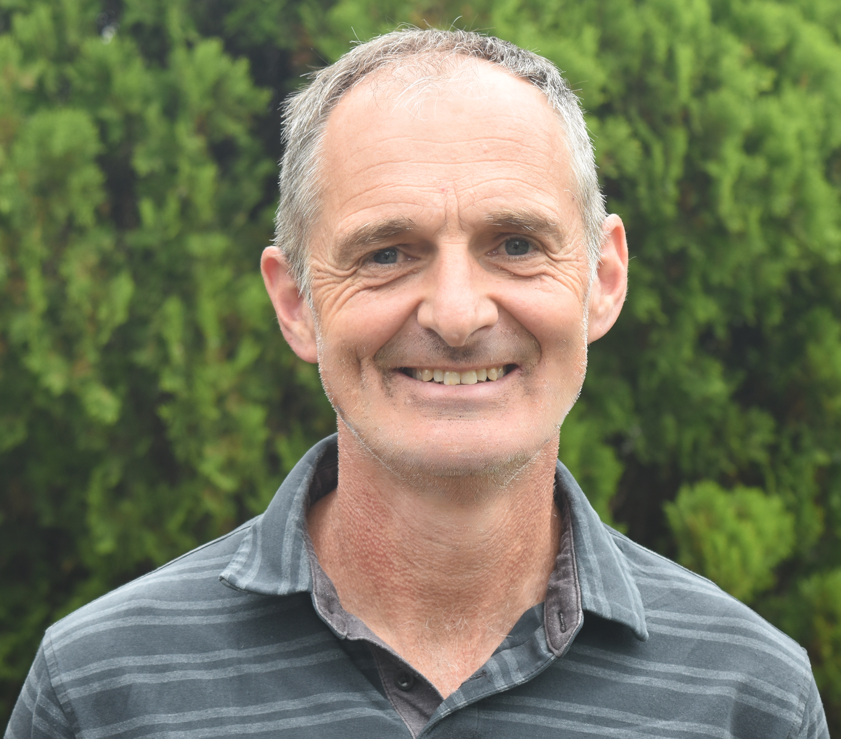 Close-up photo of a middle-aged man smiling outdoors with green trees in the background. He has short gray hair, light skin, and is wearing a gray and black striped collared shirt.