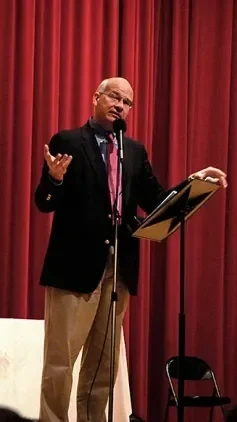 A man speaking at a microphone on stage with red curtains in the background.