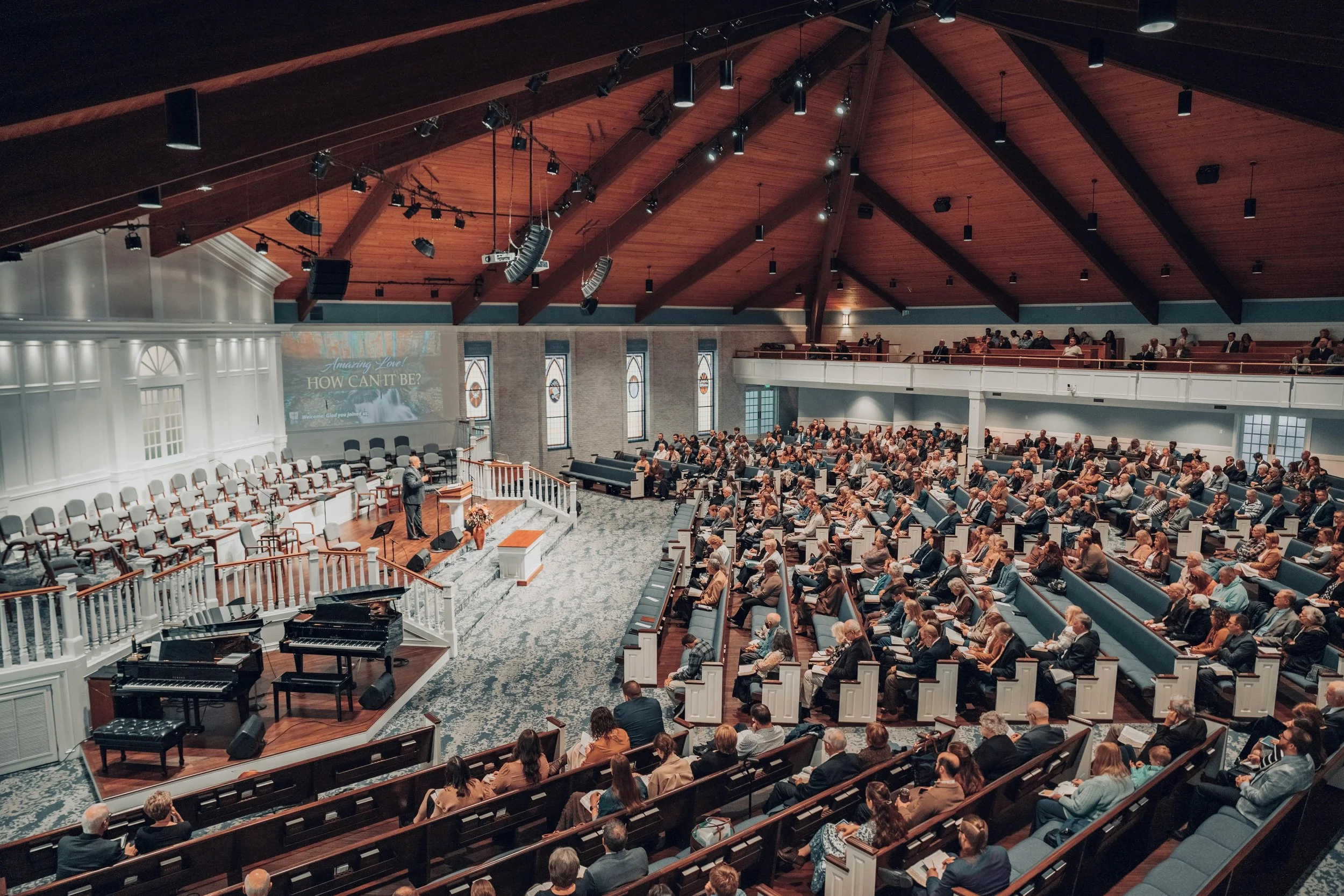 A large congregation gathers inside a church auditorium with a high wooden ceiling and stained glass windows. A man stands at a podium addressing the audience, with musical instruments including pianos in the foreground. The congregation seats are filled with people listening attentively.