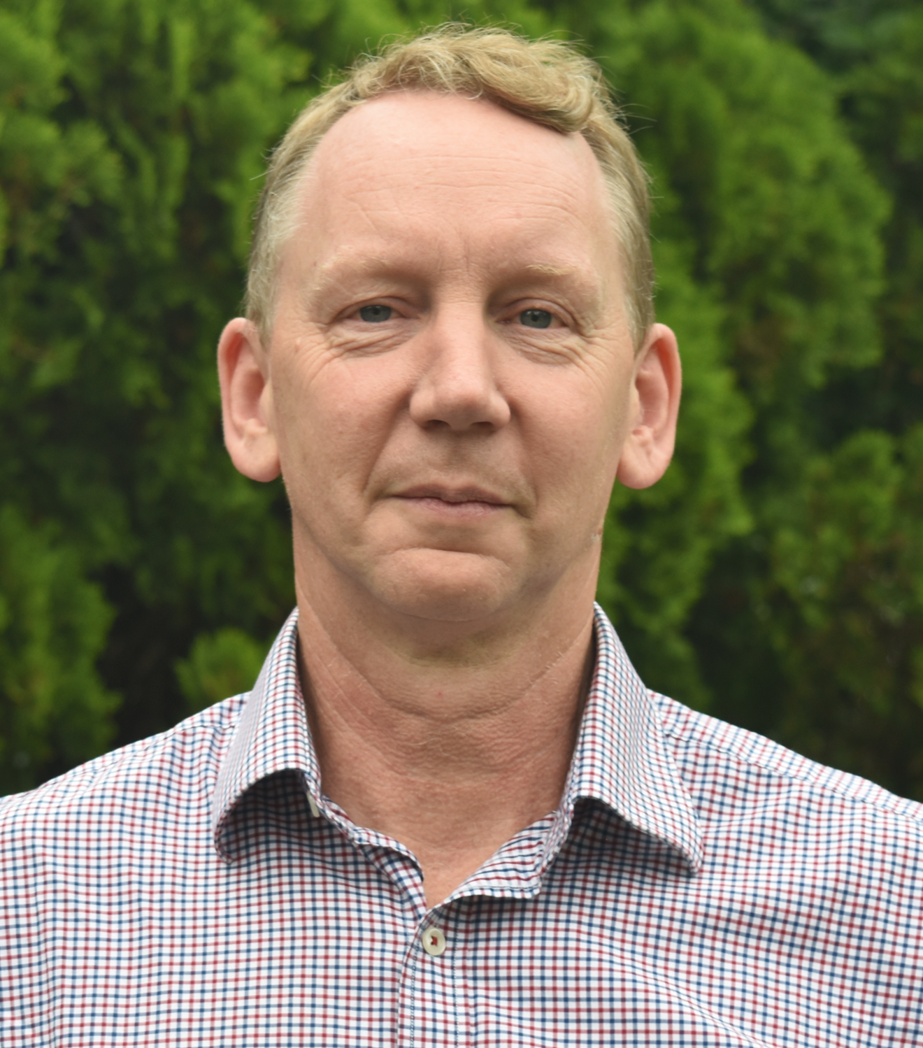 A middle-aged man with light skin and short, light brown hair, wearing a red and white checked button-up shirt, standing outdoors in front of green foliage.