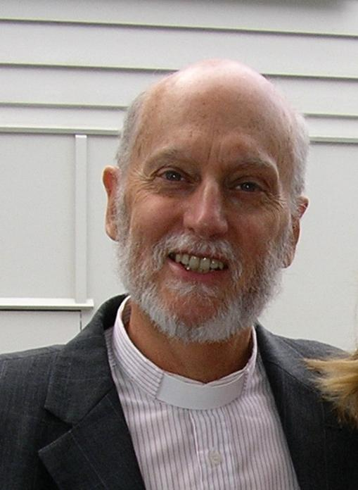 A smiling older man with a beard and balding head, wearing a suit jacket and clerical collar, standing outdoors in front of a building with horizontal siding.